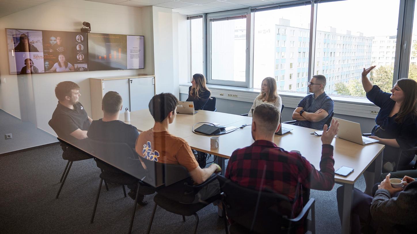 Terezia in a team meeting with colleagues around a desk and remote participants on the screen