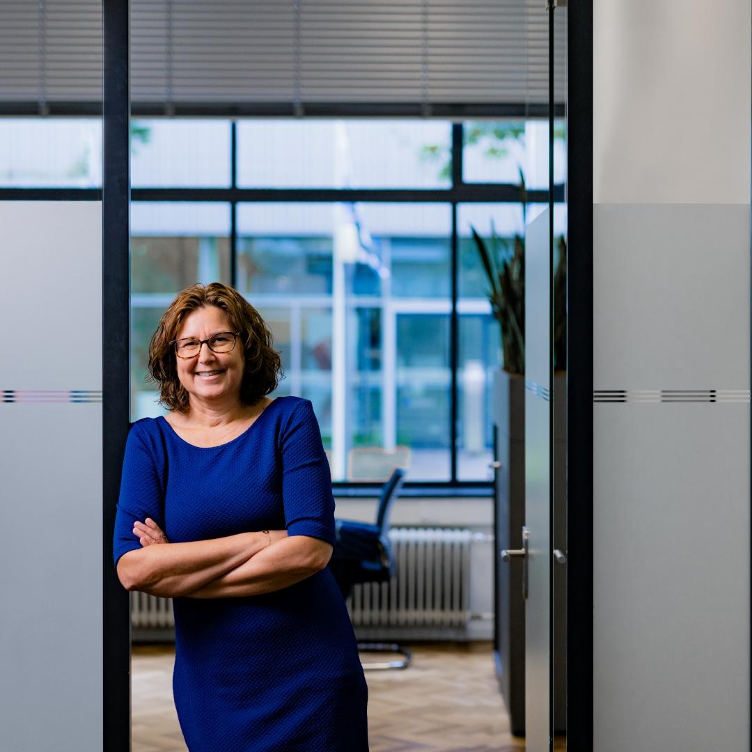 Portrait of Femke, leaning against the entry of her office, smiling at the camera