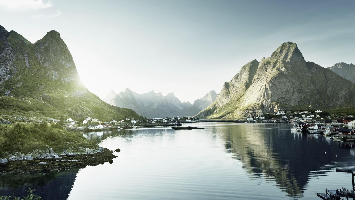 Scenic view of a fjord surrounded by steep, rocky mountains with small coastal villages and calm, reflective water under a clear sky.
