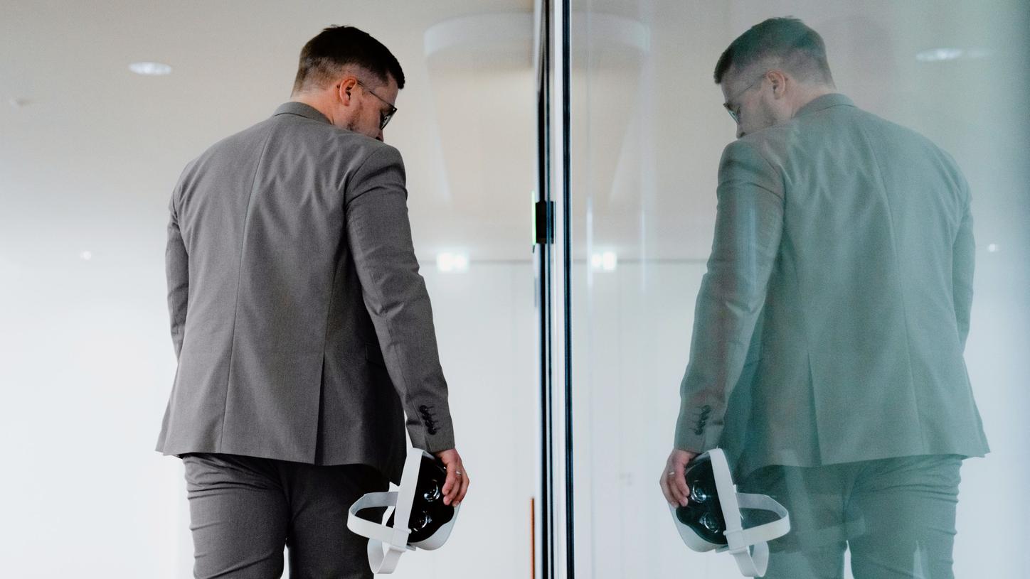 Back view of Anton, holding a pair of virtual reality glasses. Opposite is a glass facade in which he is reflected.