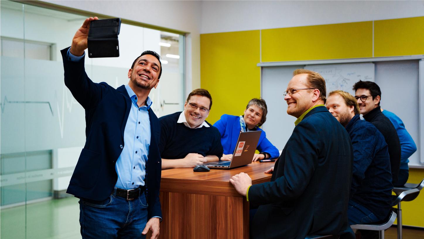 Photo in a meeting room at a table. Chiheb Dahmani takes a photo of his team with his smartphone. He and his colleagues Claudia Igney, Thorsten Gecks, Alexander Horn, Thomas Mutzke, Josef Zeidler, and Jörg Hofmann smile at the smartphone. In the background, a yellow wall with a whiteboard can be seen.