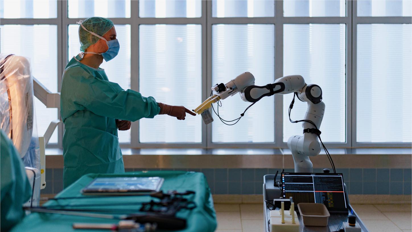 Photo in the test operating room at the Technical University of Munich hospital. A man in surgical attire with a gown, mask, cap, and gloves points to a spot in the room with his finger. The gripper of the AURORA assistant robot prototype moves towards the finger.