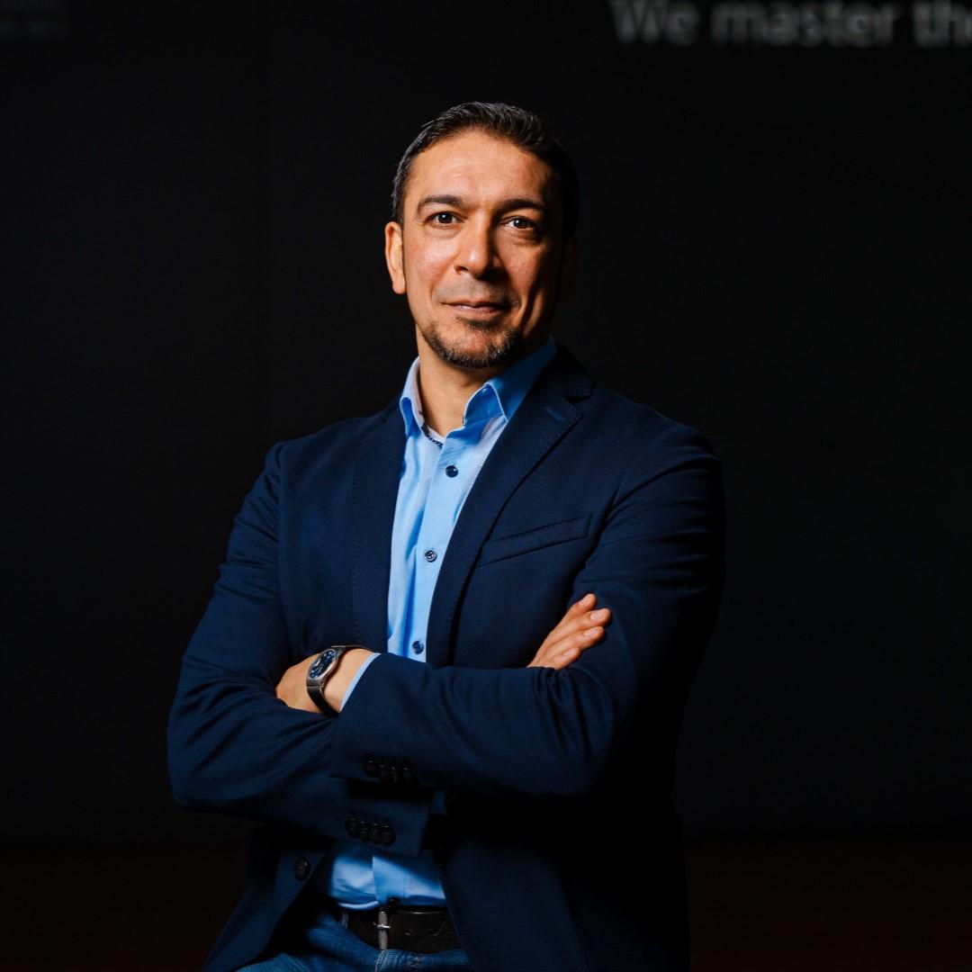 Portrait photo of Chiheb Dahmani. He stands in front of a dark background and smiles slightly towards the camera with his arms crossed.
