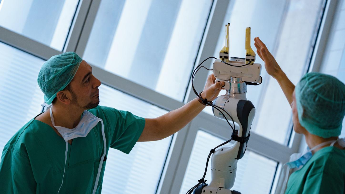 Foto von Chiheb Dahmani und Lukas Bernhard, wissenschaftlicher Leiter Forschungsgruppe für minimal-invasive interdisziplinäre therapeutische Intervention am Klinikum der Technischen Universität München. Die beiden Männer tragen OP-Kleidung und berühren mit ihren Händen den Greifarm des Prototyps des AURORA-Assistenzroboters.