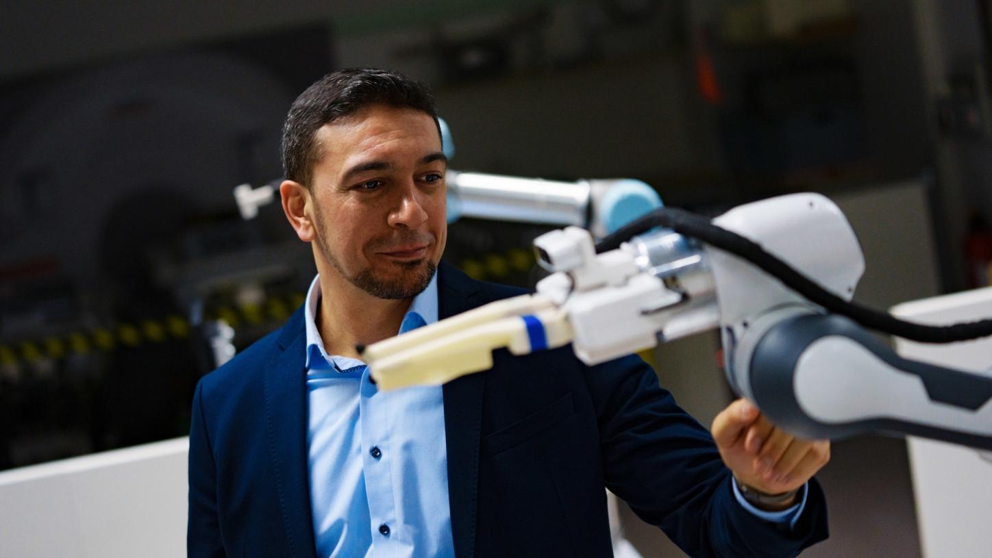 Portrait photo of Chiheb Dahmani. He is wearing a dark jacket and a light blue shirt. In the foreground, there is a robotic arm that he is lightly touching with his hand.