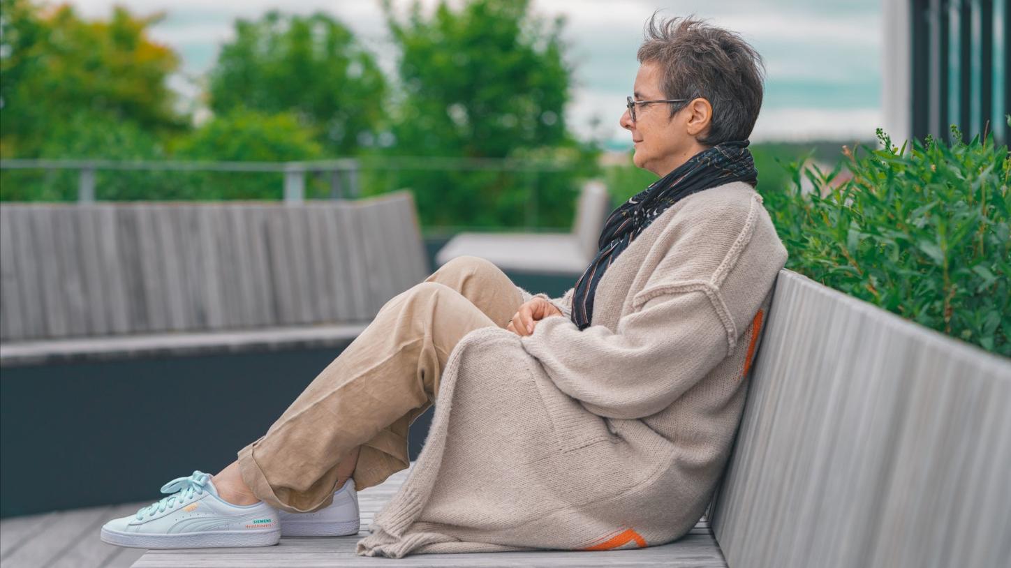 The photo shows Stefanie Vonhoff from the side in a semi-close-up shot. She is sitting on a wooden bench on a roof terrace, wearing beige trousers, a beige long cardigan and a dark scarf and gazing thoughtfully into the distance. The sky is cloudy. Trees and plants can be seen in the background.