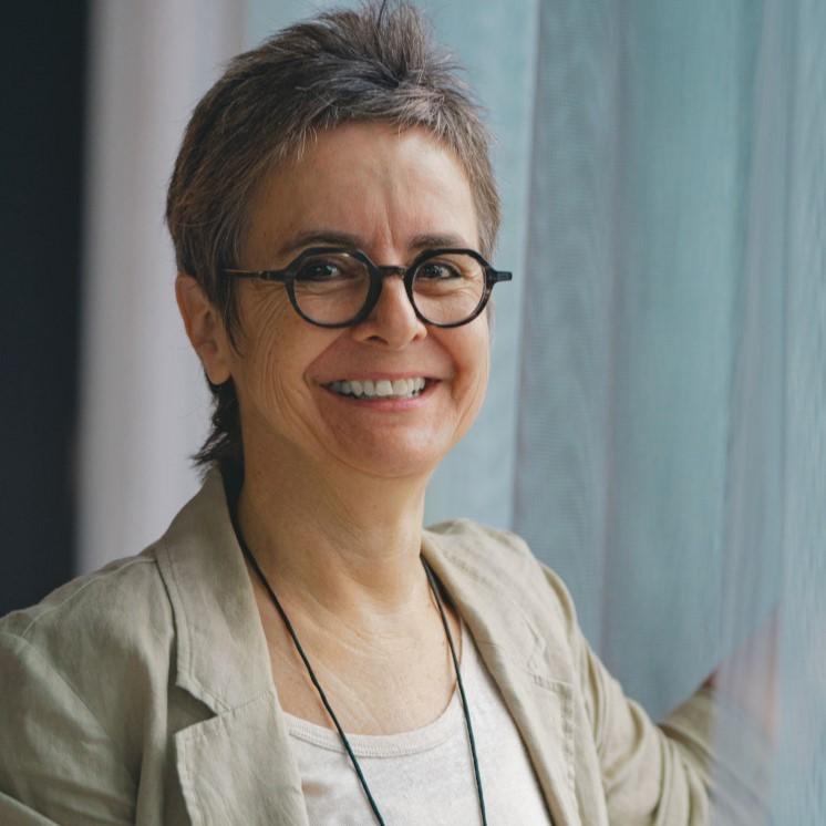 Portrait photo of Stefanie Vonhoff. She wears a beige blazer and black glasses and can be seen just up to her shoulder. She stands in front of a window with a white curtain and smiles at the camera.